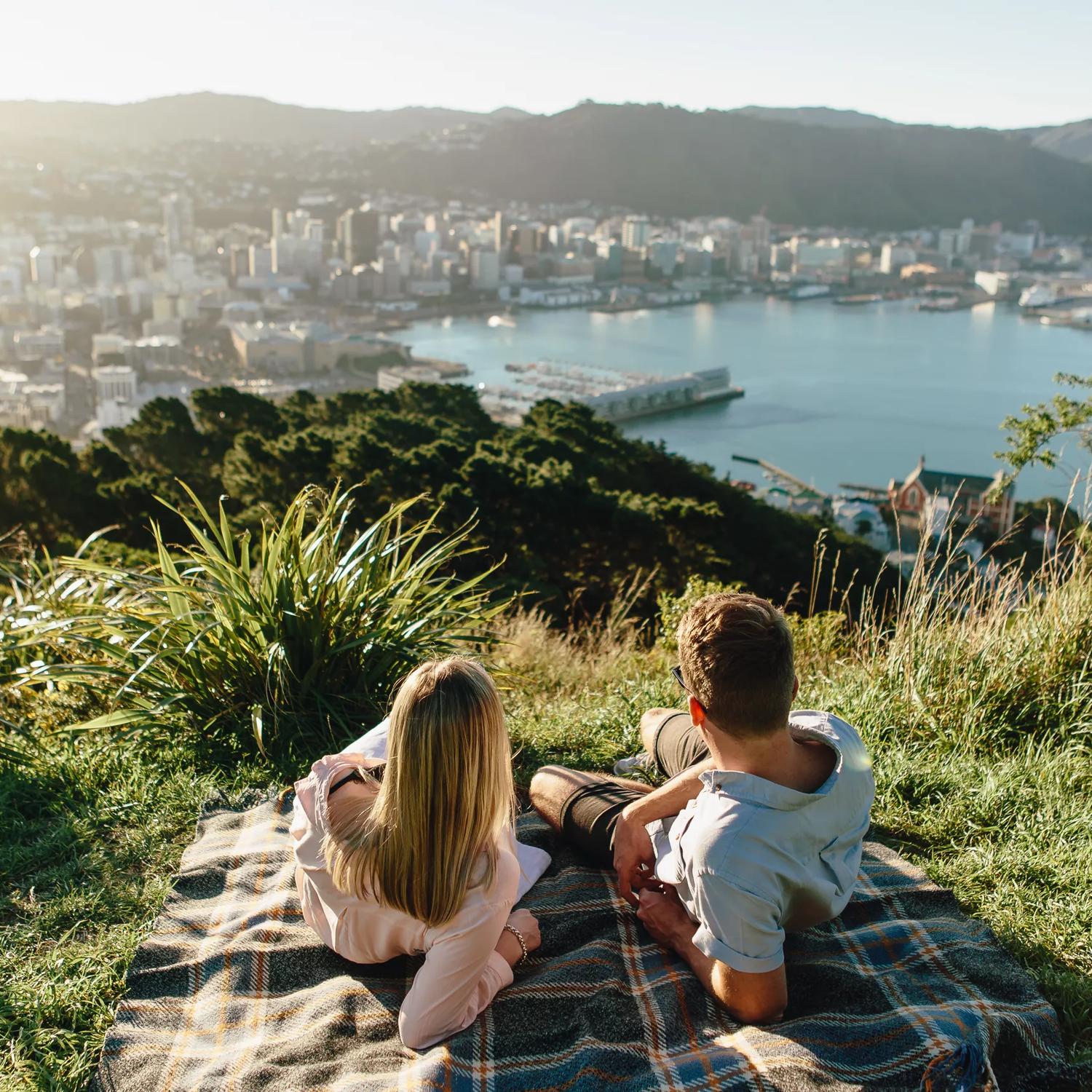 Two people lay on a plaid picnic blanket in the grass at the lookout point on Mount Victoria with a view of Wellington and the harbour int he background. 