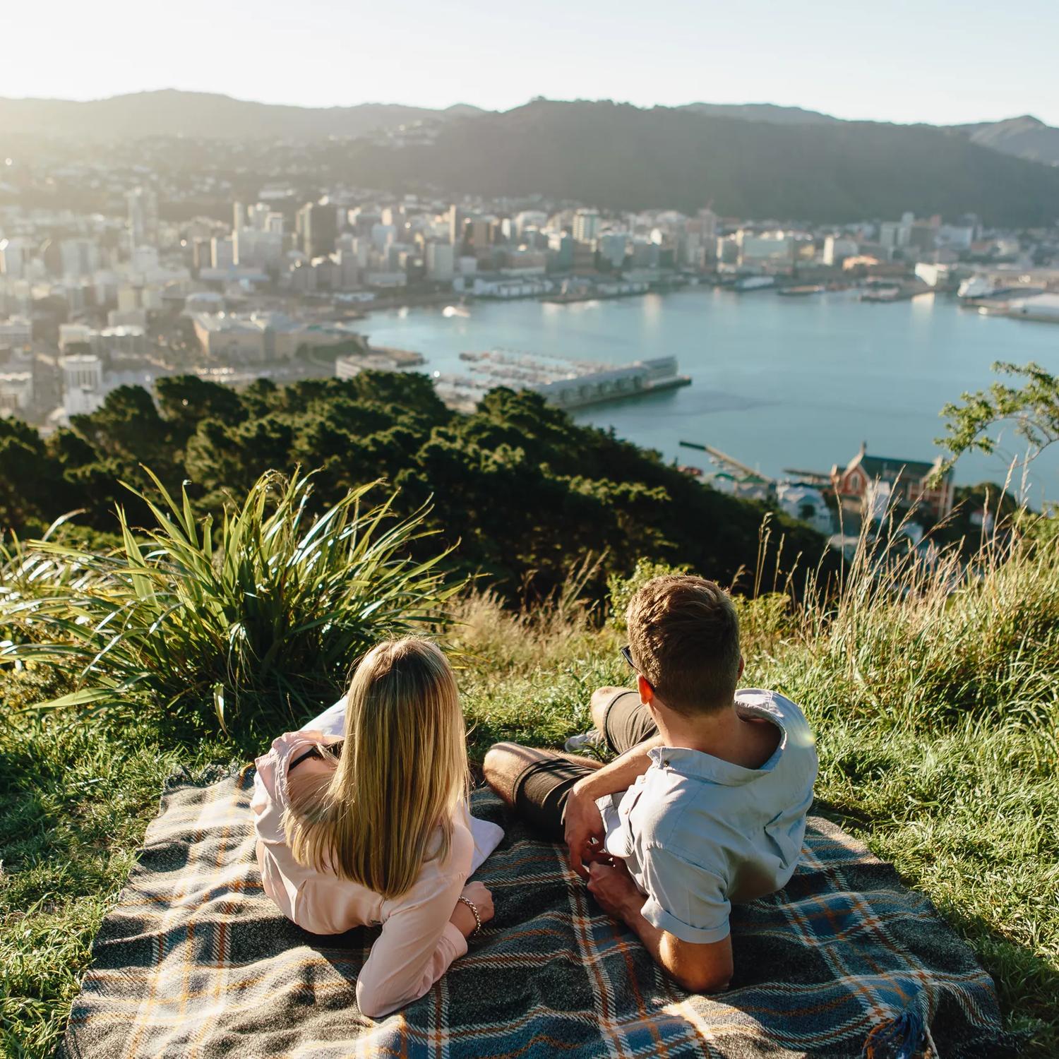 Two people lay on a plaid picnic blanket in the grass at the lookout point on Mount Victoria with a view of Wellington and the harbour int he background. 