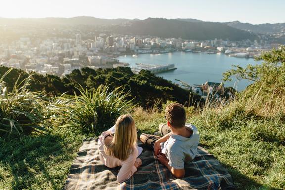 Two people lay on a plaid picnic blanket in the grass at the lookout point on Mount Victoria with a view of Wellington and the harbour int he background. 