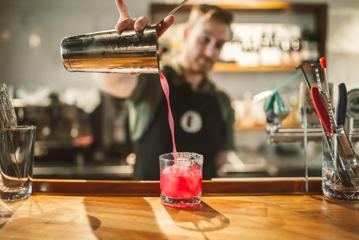 A bartender pours a pink cocktail from a shaker into a rocks glass.