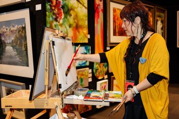 Artist weraing a yellow cardigan standing at an easel and painting with water colours.