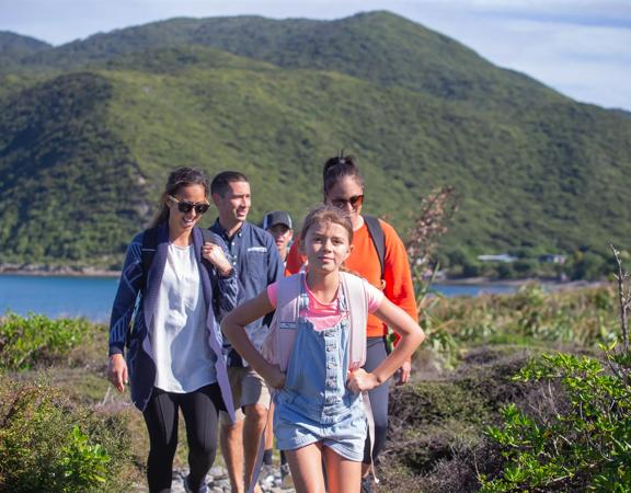 A family walks through kapiti island with green bush and blue water surrounding them.