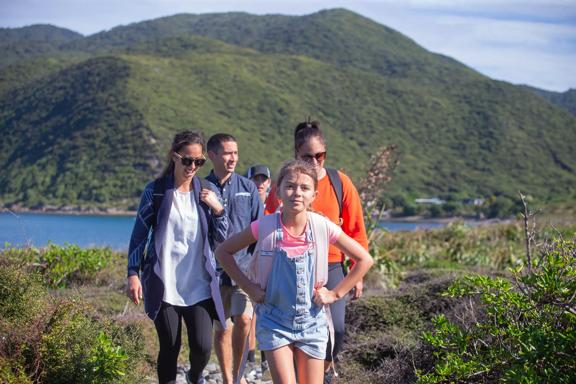 A family walks through kapiti island with green bush and blue water surrounding them.