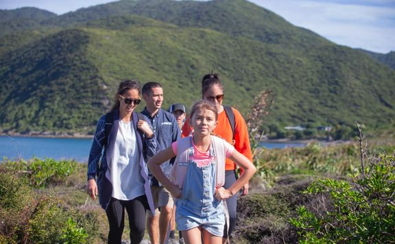 A family walks through kapiti island with green bush and blue water surrounding them.