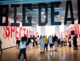 People stand in a large room inside City Gallery, looking at the art istallation which is the words 'BIG DEAL' hanging overhead.