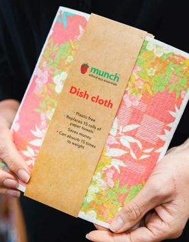 Hands hold a dishcloth. It has pink and green floral patterns and has cardboard packaging around it.