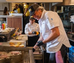 Two chefs work at a busy restaurant griddle, preparing burgers while plated buns and ingredients are arranged nearby in a kitchen setting.