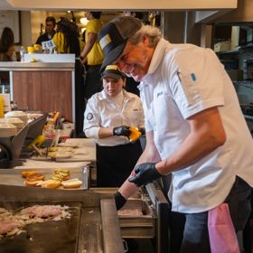 Two chefs work at a busy restaurant griddle, preparing burgers while plated buns and ingredients are arranged nearby in a kitchen setting.
