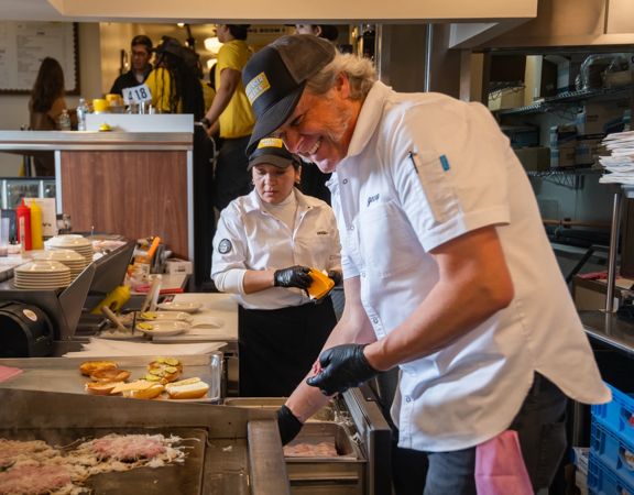 Two chefs work at a busy restaurant griddle, preparing burgers while plated buns and ingredients are arranged nearby in a kitchen setting.