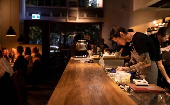 Inside Highwater Eatery, a restaurant in Te Aro, Wellington on a busy night. The dimly lit space has an open kitchen where the chefs prepare food.