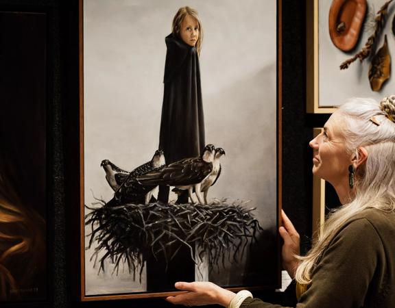 A person with long white hair wearing a dark green shirt is looking at a multi-media work of art at the NZ Art Show, a premier annual art show in Wellington.