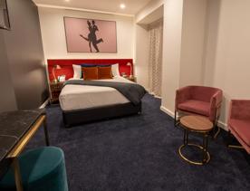 A Queen room at Tryp by Wyndham has a bed with crisp white sheets and colourful cushions against a velvet headboard. A side table and chairs are in the foreground.