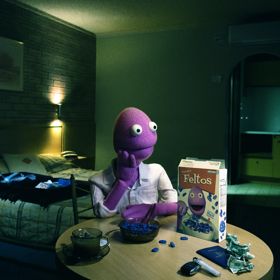 Purple puppet character seated at a table eating cereal in a dimly lit motel-style room, with a box of "Feltos" cereal displayed prominently.