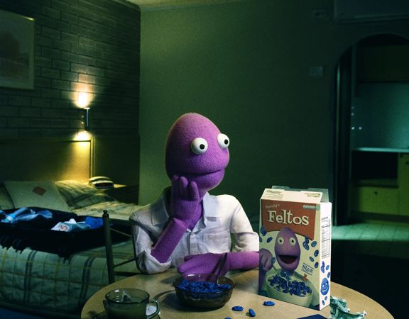Purple puppet character seated at a table eating cereal in a dimly lit motel-style room, with a box of "Feltos" cereal displayed prominently.
