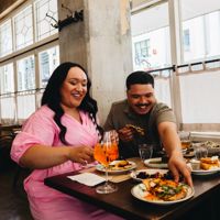 A group of three sit at a table inside Ombra near a window. One reaches across the others to grab a plate and they all laugh.