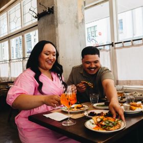 A group of three sit at a table inside Ombra near a window. One reaches across the others to grab a plate and they all laugh.