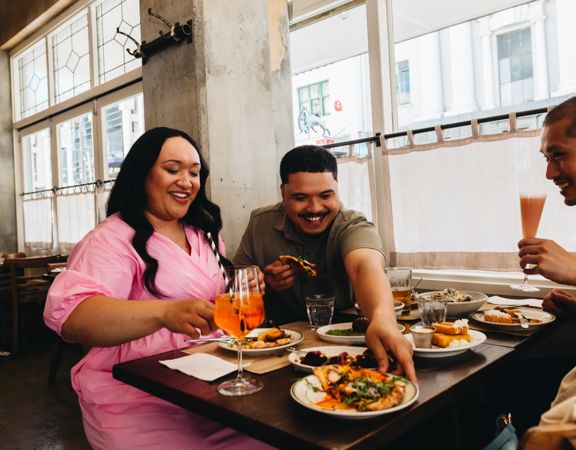 A group of three sit at a table inside Ombra near a window. One reaches across the others to grab a plate and they all laugh.