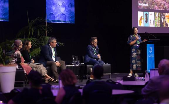 A panel discussion on stage with one person standing holding a microphone and four others seated at the Life Sciences Summit 2024 held at Tākina Wellington Convention and Exhibition Centre.
