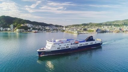 Scenic photo of Wellington harbour with blue water, blue skies, green bush and city in background, the Bluebridge ferry sits in the centre of the foreground.