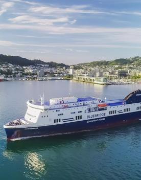 Scenic photo of Wellington harbour with blue water, blue skies, green bush and city in background, the Bluebridge ferry sits in the centre of the foreground.