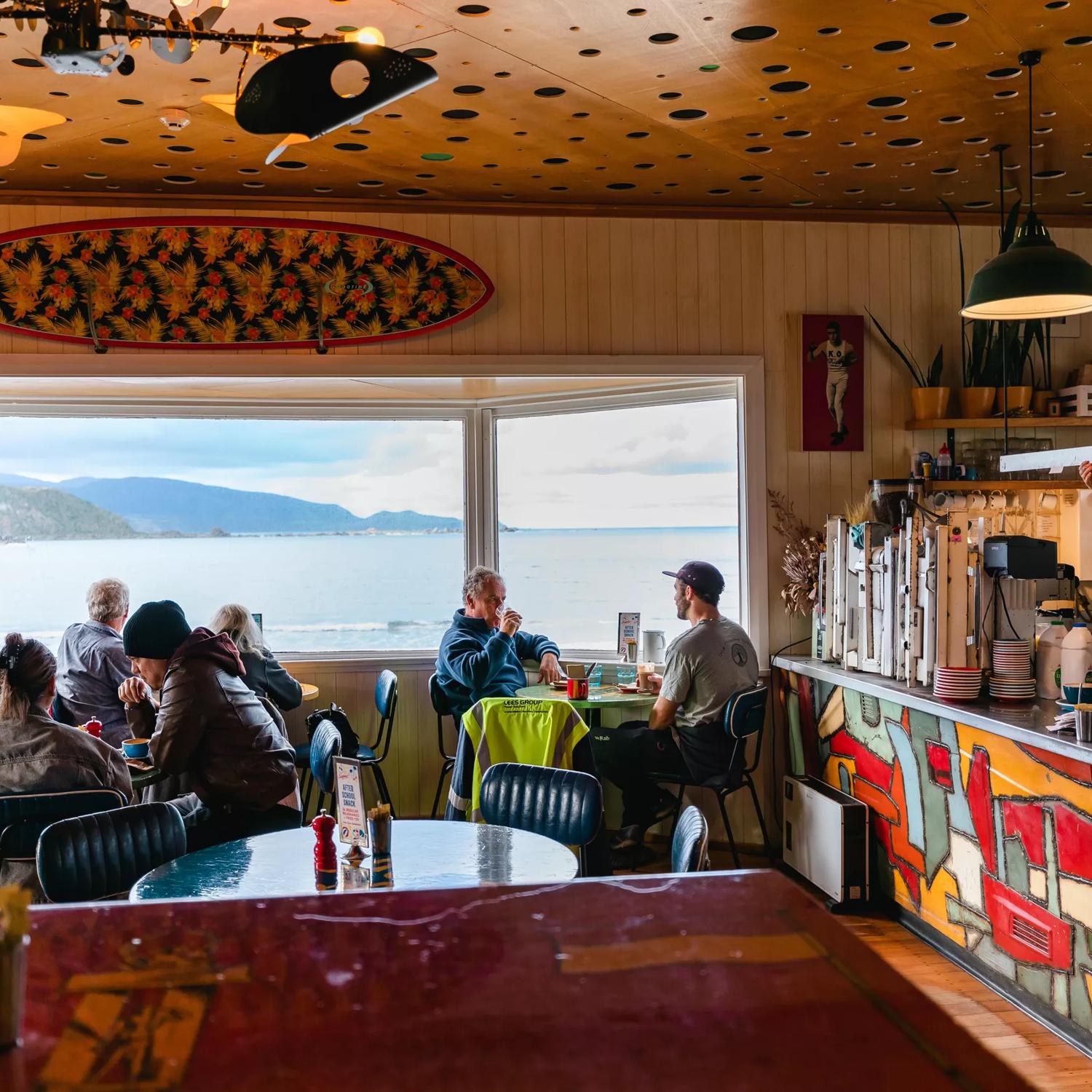 Diners sit in Maranui Cafe, overlooking Lyall Bay and Pencarrow. A barista behind the counter makes coffee.