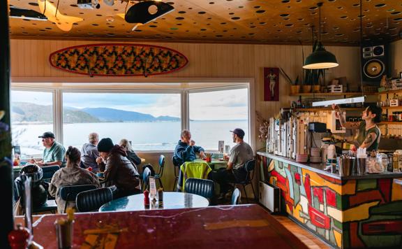 Diners sit in Maranui Cafe, overlooking Lyall Bay and Pencarrow. A barista behind the counter makes coffee.