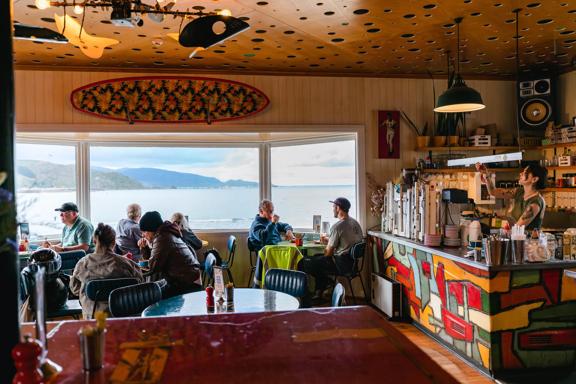 Diners sit in Maranui Cafe, overlooking Lyall Bay and Pencarrow. A barista behind the counter makes coffee.