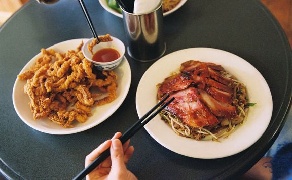 Two plates of food sit on a café table: one with sliced barbecued pork over noodles and the other with crispy fried squid and dipping sauce, as diners use chopsticks to serve themselves.