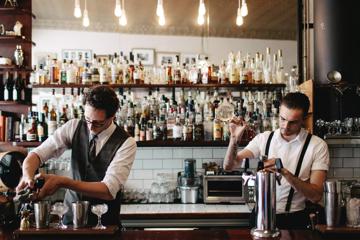 2 bartenders stand behind a bar, both pouring and mixing cocktails. A large collection of bottles sit behind them on a shelf.