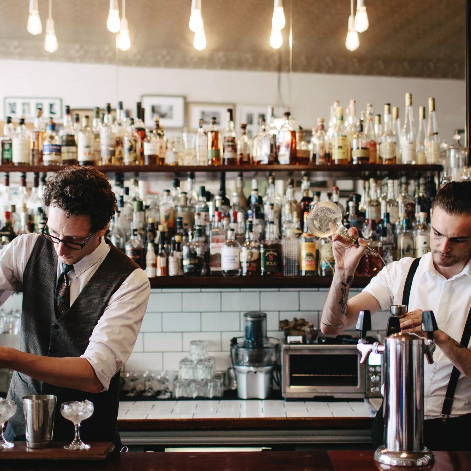 2 bartenders stand behind a bar, both pouring and mixing cocktails. A large collection of bottles sit behind them on a shelf.