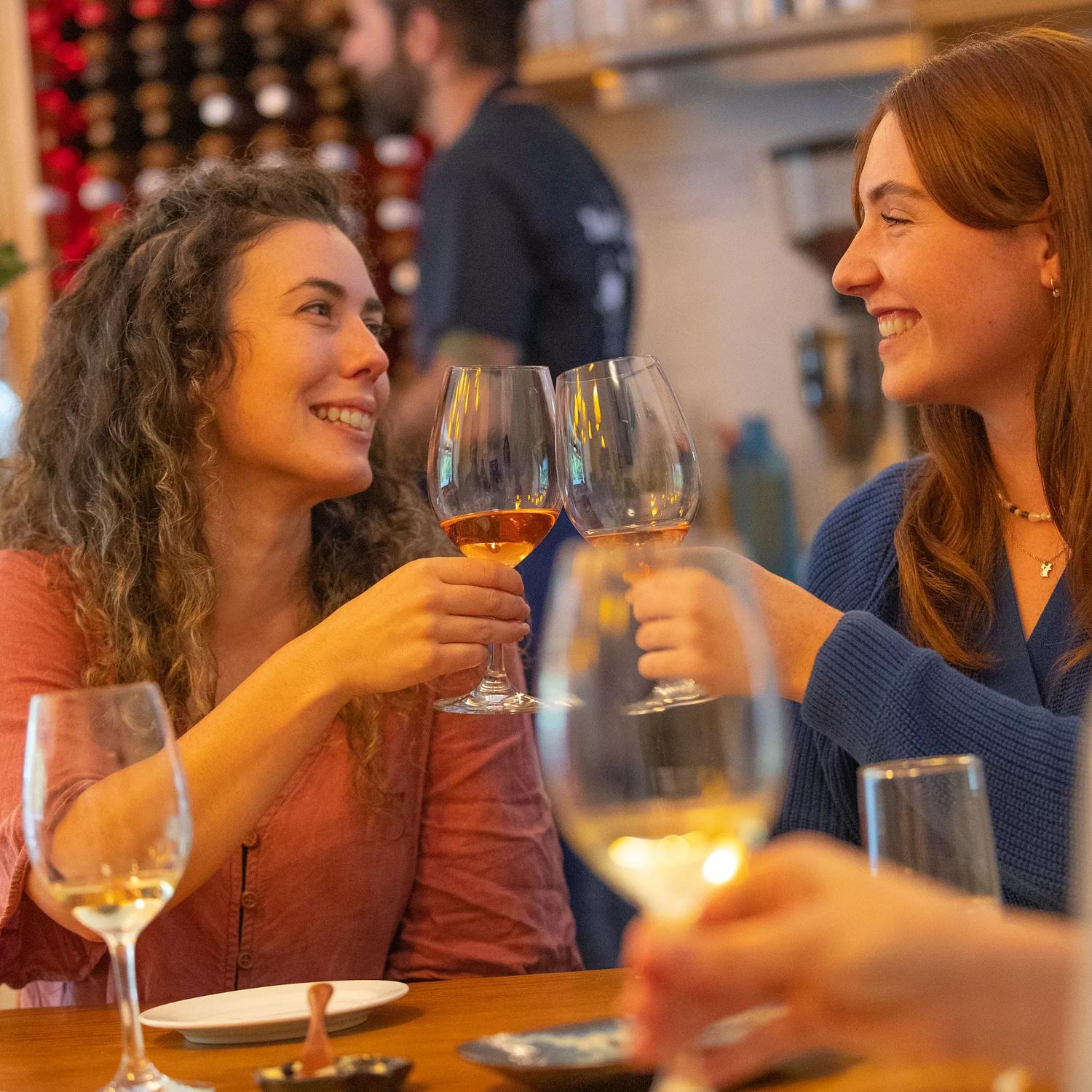 Two smiling friends clink their wine glasses while eating at a restaurant.