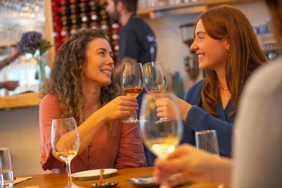 Two smiling friends clink their wine glasses while eating at a restaurant.