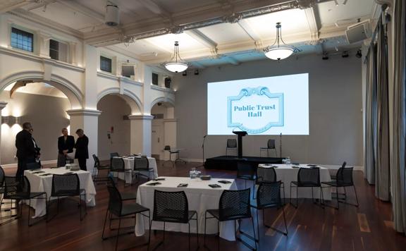 Side view of conference room with hard wood floors, 4 tables each with 4 chairs, facing towards a projector in the centre of the room and podium.