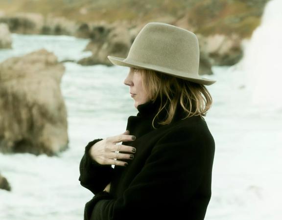 Side on photo of Beth Orton standing on a beach wearing a brown hat and a black coat.