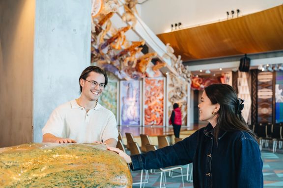 Two people touching a large rock at Museum of New Zealand Te Papa Tongarewa with the marae in the background.