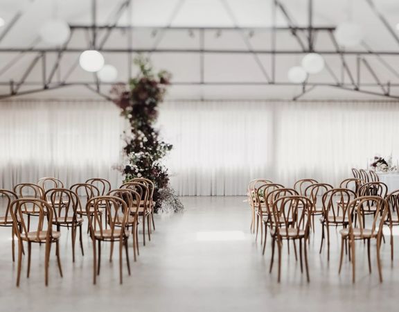 The interior of Whisky and Wood event venue with wooden chars arranged in two groups of twelve, a white curtain along the back wall, exposed brick and a tall plant with white flowers near the middle of the room.