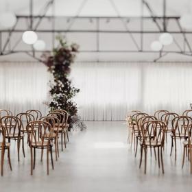 The interior of Whisky and Wood event venue with wooden chars arranged in two groups of twelve, a white curtain along the back wall, exposed brick and a tall plant with white flowers near the middle of the room. 