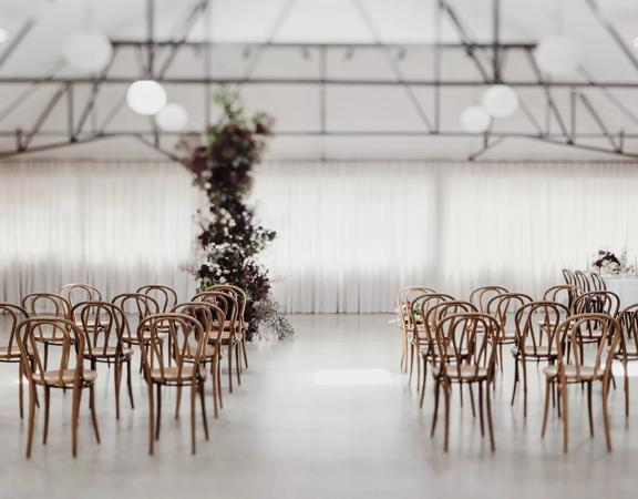 The interior of Whisky and Wood event venue with wooden chars arranged in two groups of twelve, a white curtain along the back wall, exposed brick and a tall plant with white flowers near the middle of the room. 