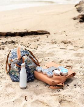 A navy blue bag with flowers and leather straps on the left, a light beige leather purse on the right, and a white water bottle in the foreground on a sandy beach. There are six tins of Seasick Sunscreen and a smartphone arranged on the light beige bag. 