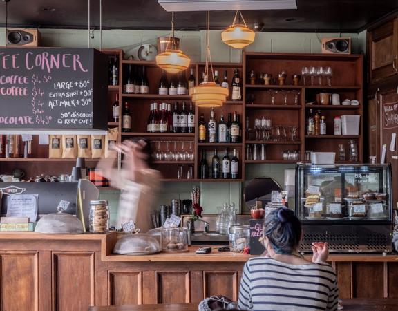 The interior of Raumati social club, customers sitting at tables and the bar staff making drinks behind the bar. A shelf of wine and spirit bottles is seen behind the bar.