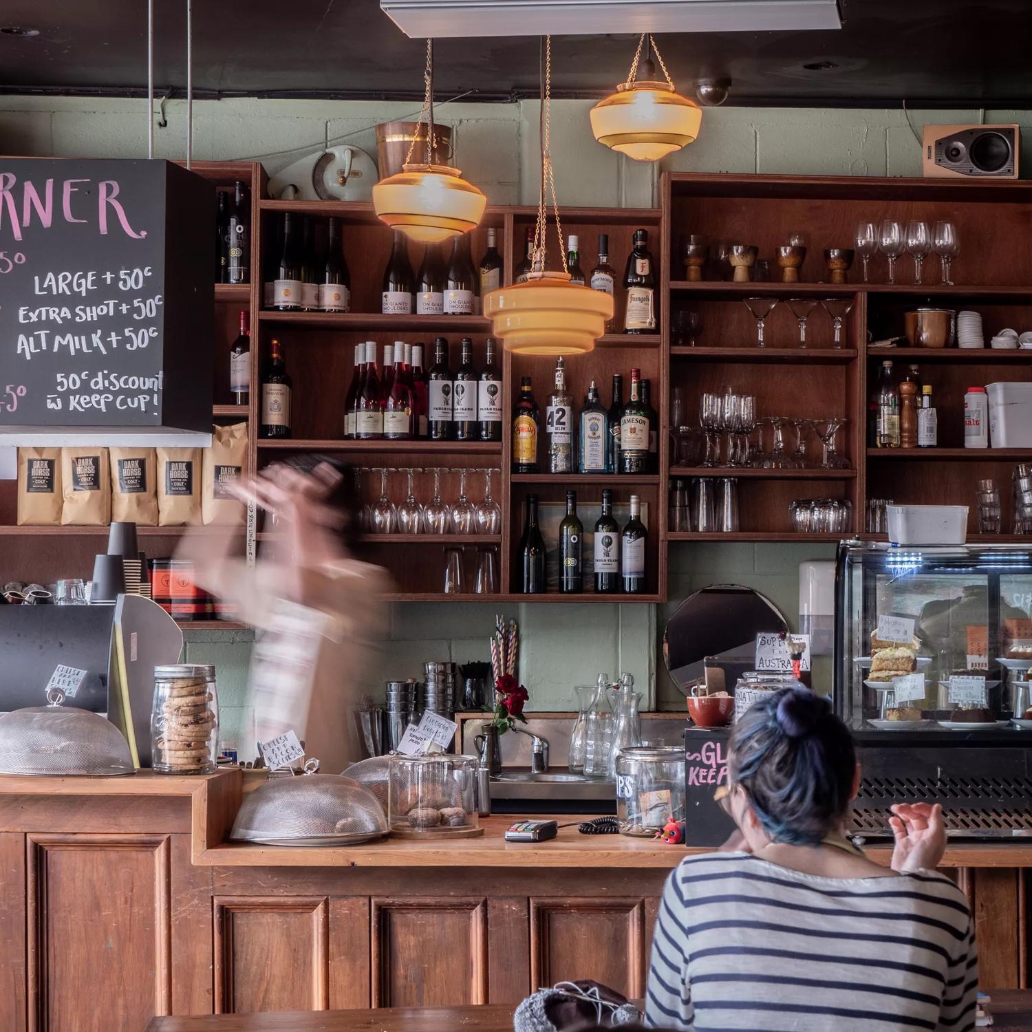 The interior of Raumati social club, customers sitting at tables and the bar staff making drinks behind the bar. A shelf of wine and spirit bottles is seen behind the bar.