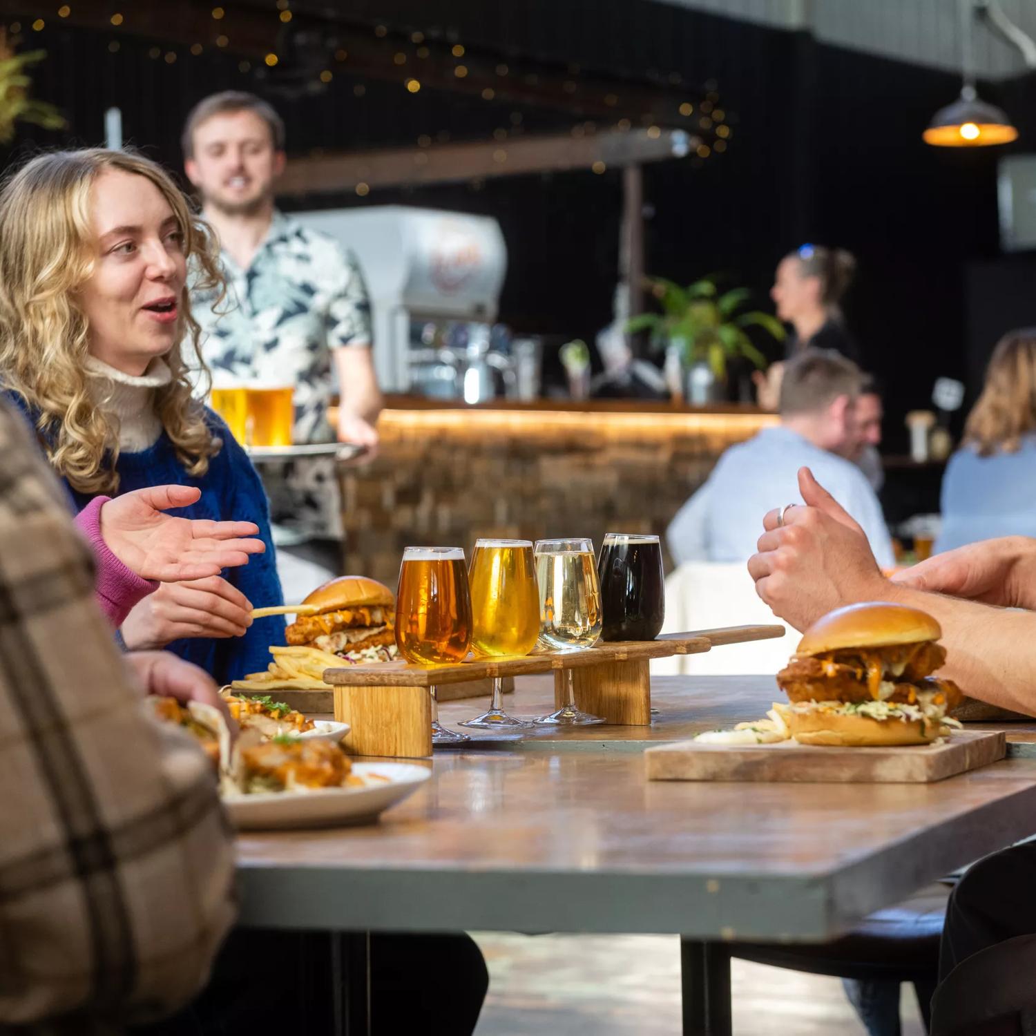 People sitting at a table with beers and burgers at Aro Bar in Brewtown, Upper Hutt. The main focus point is a person talking.