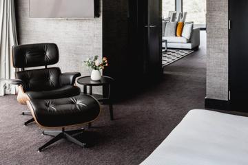 The interior of the Residence Master Suite Master Bedroom at the Bolton Hotel in Wellington with grey textured wallpaper, a modern black leather chair matching ottoman and small black circular side table.