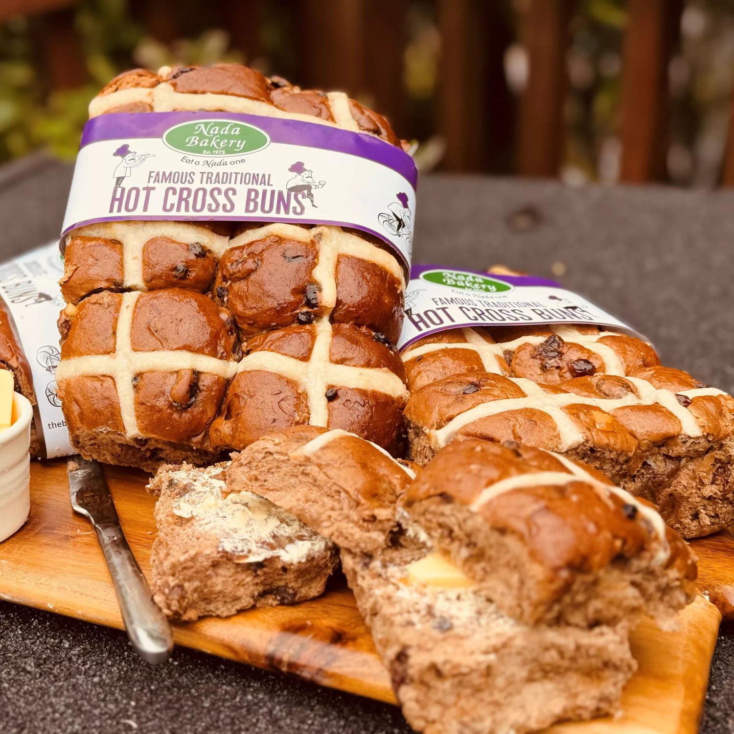 A wooden tray laden with multiple hot cross buns. Some are still in a six-pack formation and have marketing labels attached. To the left lies a knife and a small bowl of butter.