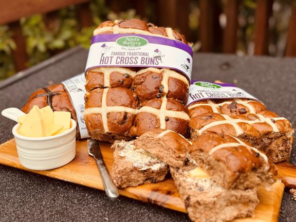 A wooden tray laden with multiple hot cross buns. Some are still in a six-pack formation and have marketing labels attached. To the left lies a knife and a small bowl of butter.