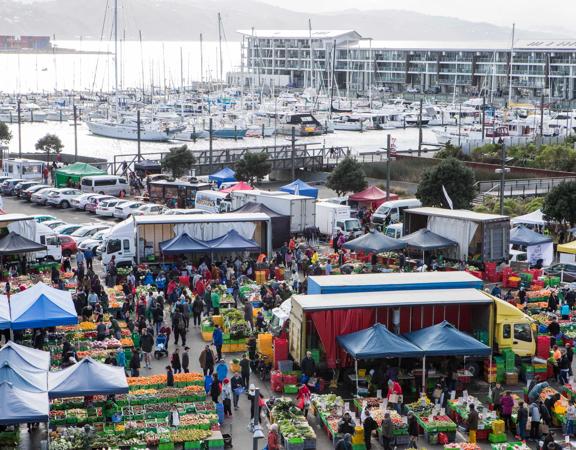 An aerial view of the bustling Habourside Market in Te Aro Wellington. There are tents set up, heaps of colourful produce and many people shopping. 