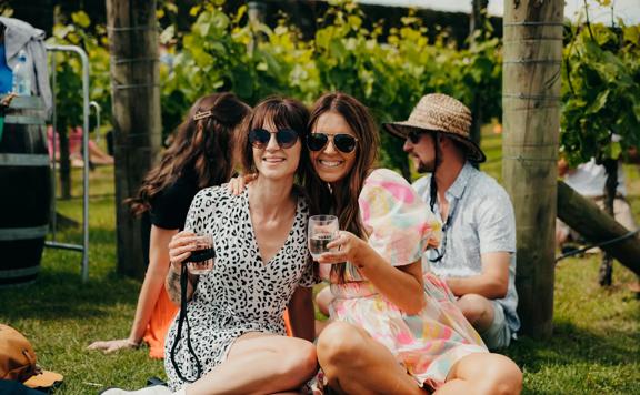 Two friends sit on a lawn drinking wine.