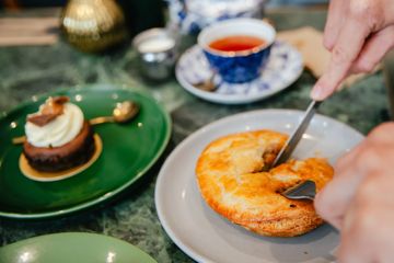 A person cuts into a glazed pastry on a plate at Uncle Leo's Bakery in Hataitai, with a small cake and a cup of tea on the table nearby.