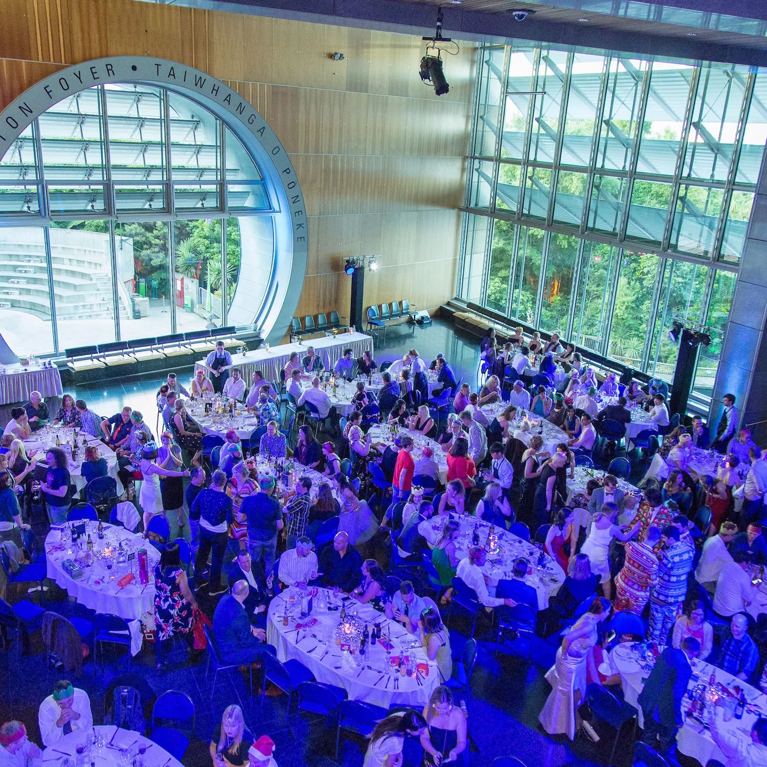 Inside the Wellington Foyer, Taiwhanga o Pōneke, during an event. There are many people gathered around tables with purple-coloured lighting.