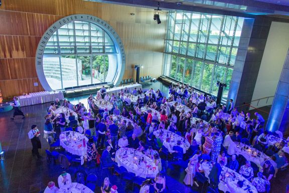 Inside the Wellington Foyer, Taiwhanga o Pōneke, during an event. There are many people gathered around tables with purple-coloured lighting.